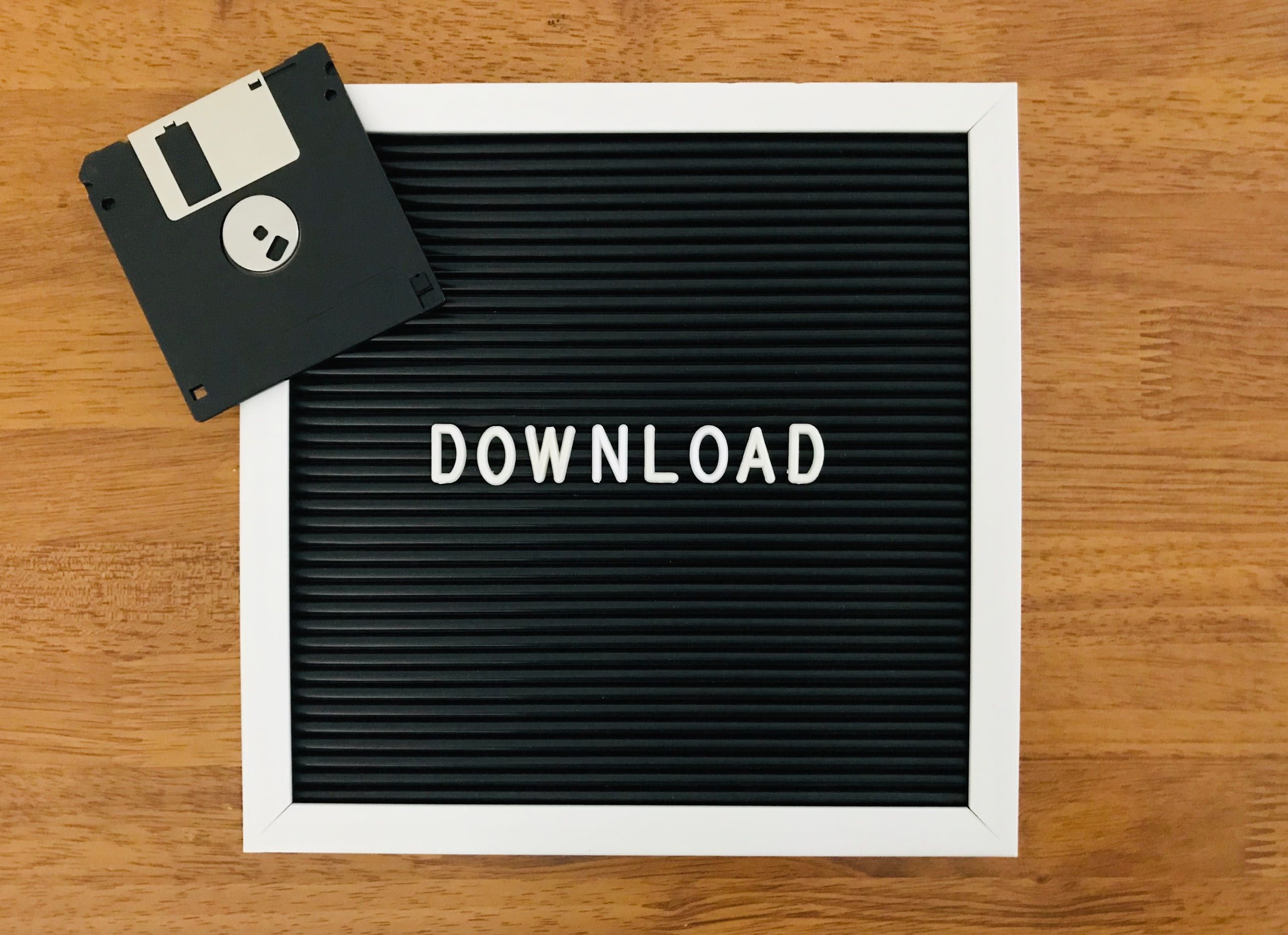 A black felt letter board spelling DOWNLOAD, a 3.5-inch floppy disk propped on its corner, on a wooden table.